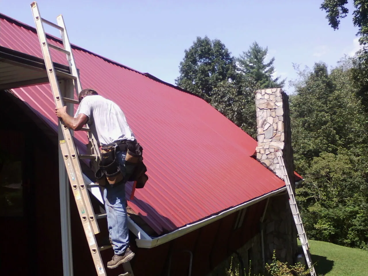 Roofer on a ladder beside a red metal roof and stone chimney — Soffit & Fascia Repair by Roofing Brothers in Marmora
